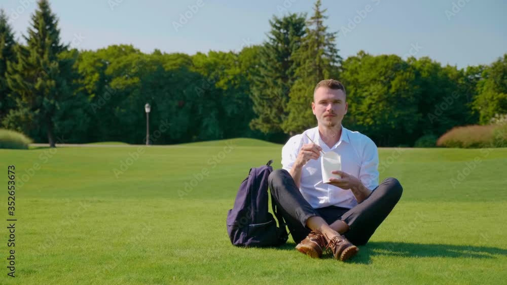 Man looks at the camera with a vegetarian lunch in an ecologic package in his hand. Guy is sitting in the park on the green grass, holding a paper package in his hand with a salad and wooden sticks Man looks at the camera with a vegetarian lunch in an ecologic package in his hand. Guy is sitting in the park on the green grass, holding a paper package in his hand with a salad and wooden sticks