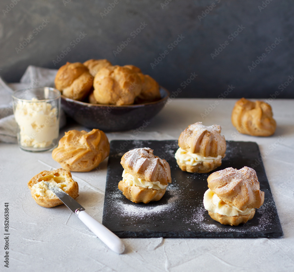 Cream puffs or profiteroles with whipped cream dusted with sugar powder on slate board, filling split puff with cream using knife