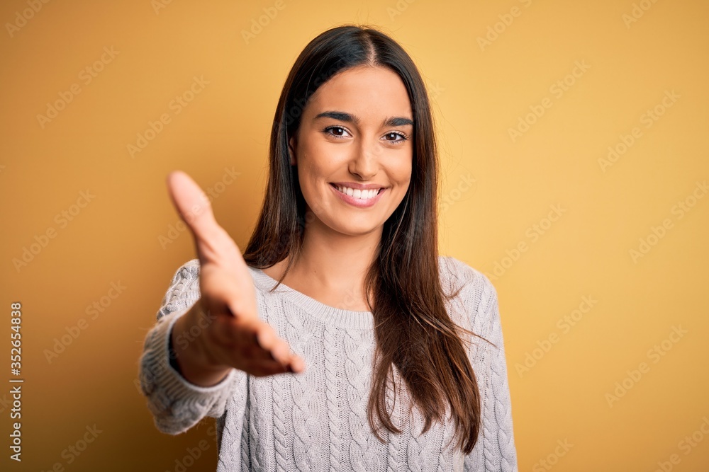 Young beautiful brunette woman wearing white casual sweater over yellow background smiling friendly offering handshake as greeting and welcoming. Successful business. Young beautiful brunette woman wearing white casual sweater over yellow background smiling friendly offering handshake as greeting and welcoming. Successful business.