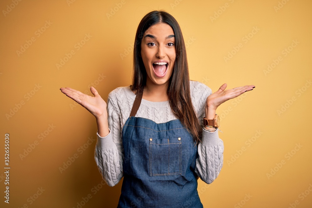Young beautiful baker woman wearing apron uniform cooking over yellow background celebrating crazy and amazed for success with arms raised and open eyes screaming excited. Winner concept Young beautiful baker woman wearing apron uniform cooking over yellow background celebrating crazy and amazed for success with arms raised and open eyes screaming excited. Winner concept