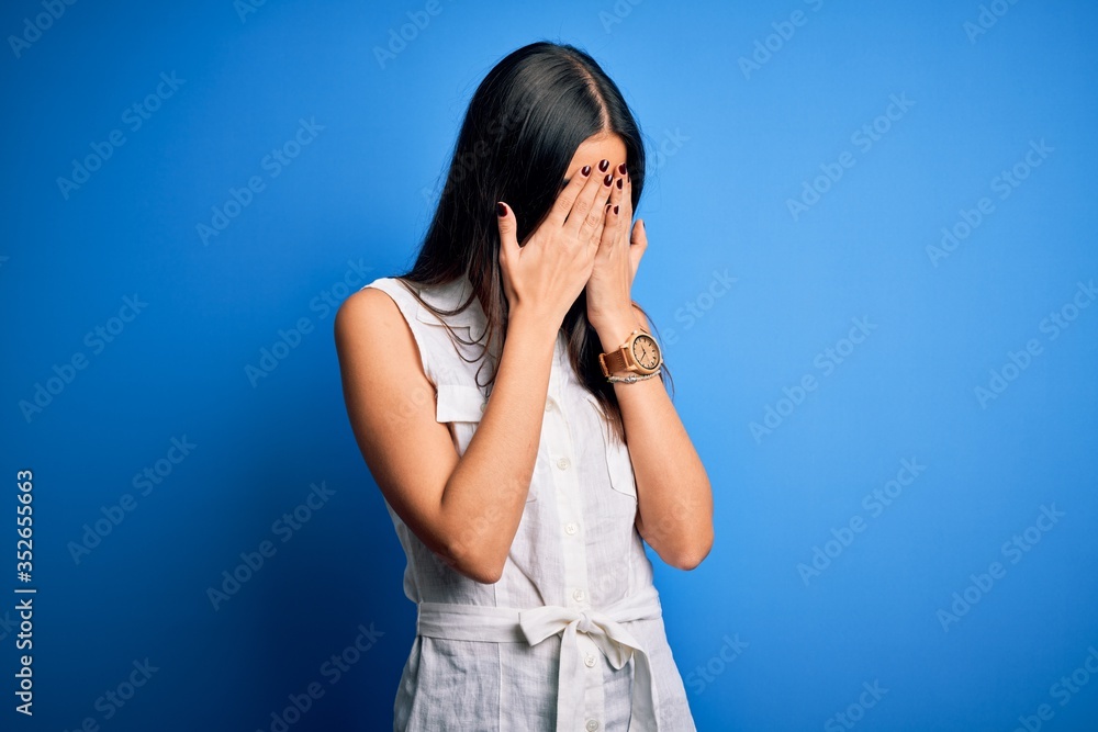 Young beautiful brunette woman wearing casual dress over isolated blue background with sad expression covering face with hands while crying. Depression concept. Young beautiful brunette woman wearing casual dress over isolated blue background with sad expression covering face with hands while crying. Depression concept.