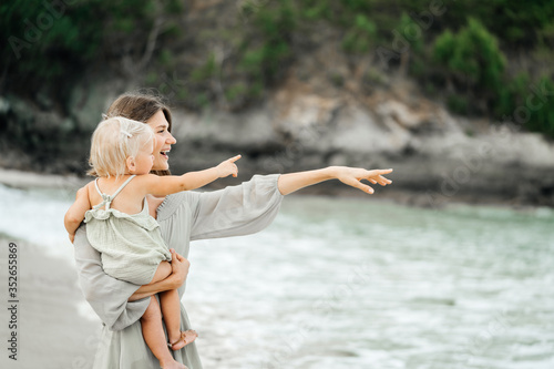 Young mother and her blond child are resting on a tropical beach showing with hands to the horizon. Travel and motherhood.