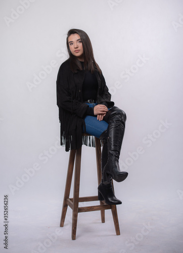 Happy beautiful girl model in black dress posing in studio isolated on white background,Teen girl in in blue jeans and black boots Full length portrait,sitting on a chair