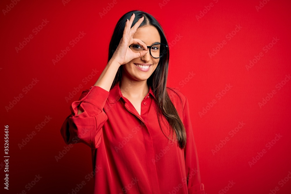 Young beautiful brunette woman wearing casual shirt and glasses over red background doing ok gesture with hand smiling, eye looking through fingers with happy face.