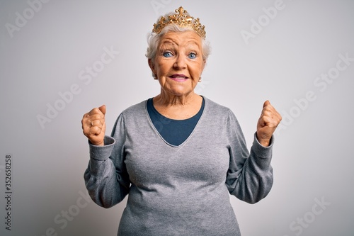 Senior beautiful grey-haired woman wearing golden queen crown over white background celebrating surprised and amazed for success with arms raised and open eyes. Winner concept.