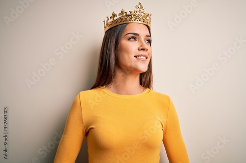 Young beautiful brunette woman wearing golden queen crown over white background looking away to side with smile on face, natural expression. Laughing confident. photo