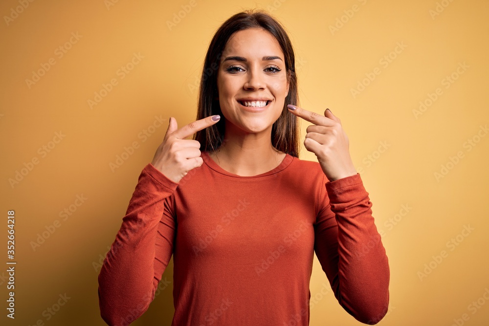 Young beautiful brunette woman wearing casual t-shirt standing over yellow background smiling cheerful showing and pointing with fingers teeth and mouth. Dental health concept. Young beautiful brunette woman wearing casual t-shirt standing over yellow background smiling cheerful showing and pointing with fingers teeth and mouth. Dental health concept.