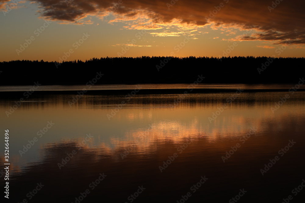 Mirror image of pink light sky with bright burning crimson clouds in the lake at dawn.The illuminated black forest on the horizon is copied in the calm water.A glossy picture in the twilight.Russia