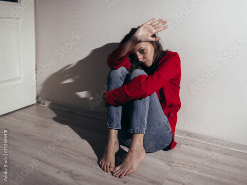 A girl in a red shirt and jeans sits sad against the wall. Domestic violence. She raised her hand asking for a stop.