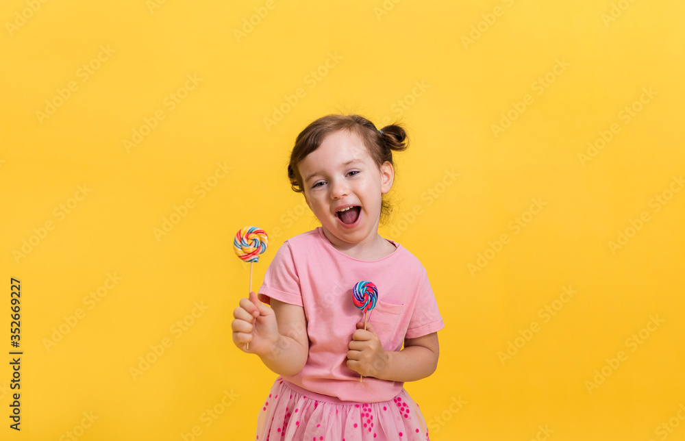 A laughing little girl in a pink t-shirt with ponytails holds two colorful lollipops on a yellow isolated background with space for text. A laughing little girl in a pink t-shirt with ponytails holds two colorful lollipops on a yellow isolated background with space for text.