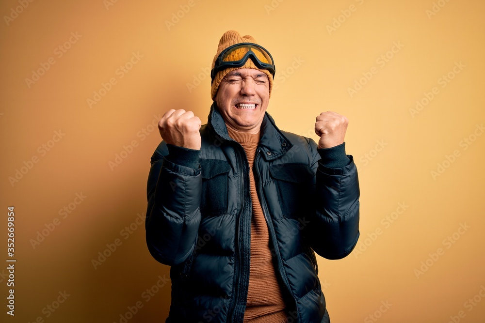 Middle age handsome grey-haired skier man on vacation wearing ski goggles very happy and excited doing winner gesture with arms raised, smiling and screaming for success. Celebration concept. Middle age handsome grey-haired skier man on vacation wearing ski goggles very happy and excited doing winner gesture with arms raised, smiling and screaming for success. Celebration concept.
