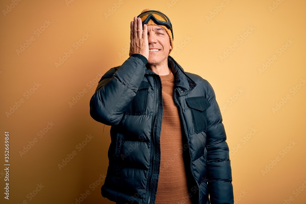 Middle age handsome grey-haired skier man on vacation wearing ski goggles covering one eye with hand, confident smile on face and surprise emotion.