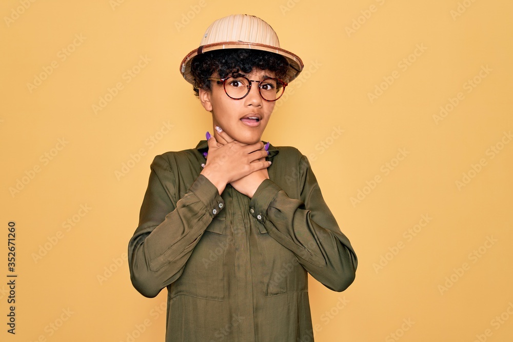 Young beautiful african american tourist woman wearing glasses and explorer hat shouting and suffocate because painful strangle. Health problem. Asphyxiate and suicide concept. Young beautiful african american tourist woman wearing glasses and explorer hat shouting and suffocate because painful strangle. Health problem. Asphyxiate and suicide concept.