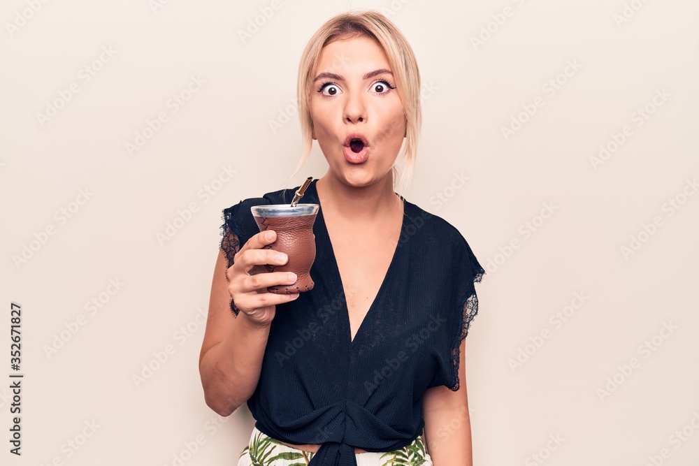Young beautiful blonde woman drinking cup of mate infusion beverage over white background scared and amazed with open mouth for surprise, disbelief face Young beautiful blonde woman drinking cup of mate infusion beverage over white background scared and amazed with open mouth for surprise, disbelief face