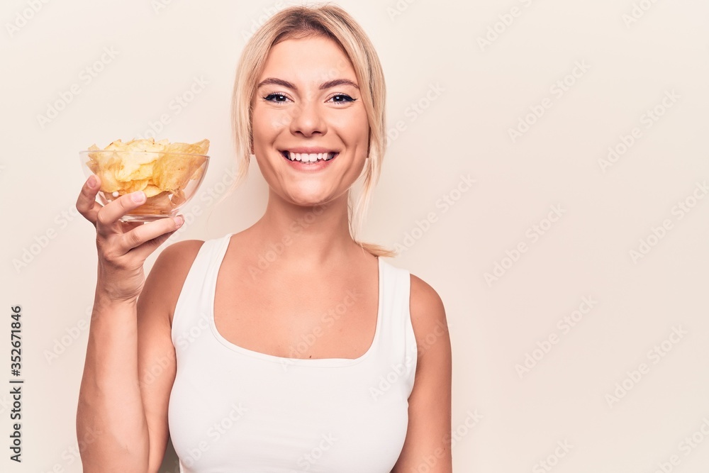 Young beautiful blonde woman holding bowl with potatoes chips over isolated white background looking positive and happy standing and smiling with a confident smile showing teeth Young beautiful blonde woman holding bowl with potatoes chips over isolated white background looking positive and happy standing and smiling with a confident smile showing teeth