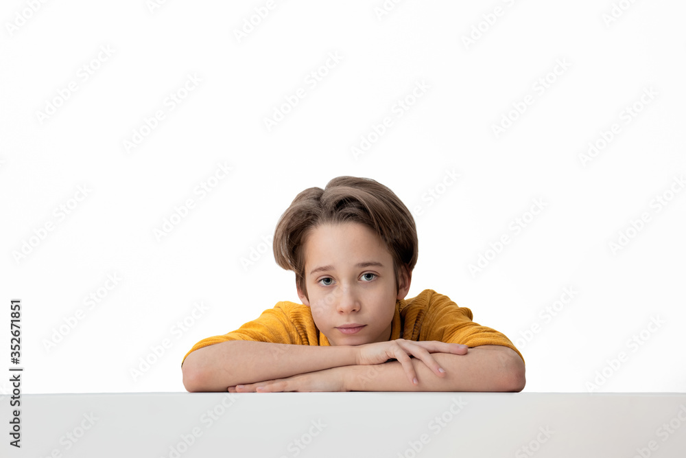 Portrait of a teenager, a schoolboy sitting at a table on a white background, looking at the camera. Adorable child, studio shot, over white background, with copy space. Portrait of a teenager, a schoolboy sitting at a table on a white background, looking at the camera. Adorable child, studio shot, over white background, with copy space.