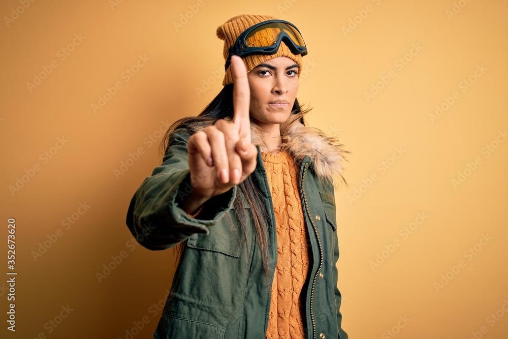 Young beautiful hispanic woman wearing ski glasses and coat for winter weather Pointing with finger up and angry expression, showing no gesture Young beautiful hispanic woman wearing ski glasses and coat for winter weather Pointing with finger up and angry expression, showing no gesture