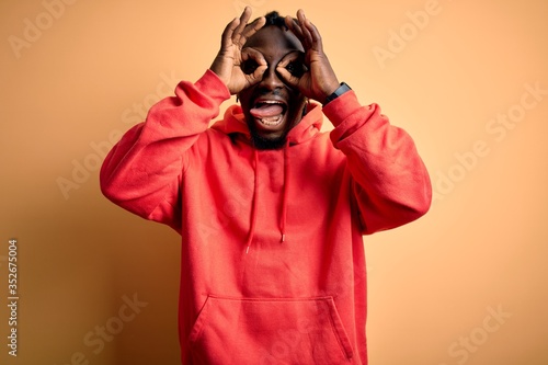 Young african american sporty man wearing sweatshirt with hoodie over yellow background doing ok gesture like binoculars sticking tongue out, eyes looking through fingers. Crazy expression. © Krakenimages.com