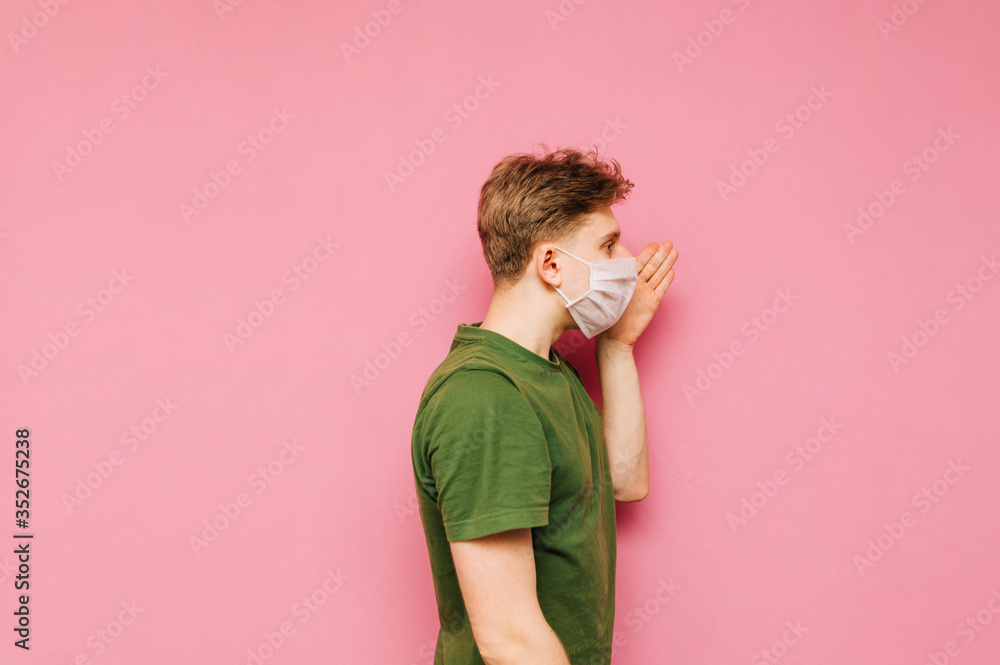 Young man in a medical mask shouts to the side with his hand to his face on a pink background. Guy in the protective white mask is talking. Coronavirus pandemic. COVID-19. Quarantine. Young man in a medical mask shouts to the side with his hand to his face on a pink background. Guy in the protective white mask is talking. Coronavirus pandemic. COVID-19. Quarantine.