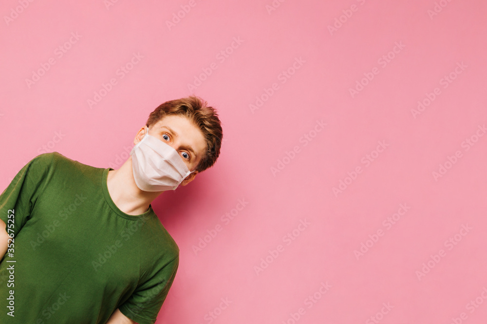 Φωτογραφία Funny guy in bright casual clothes and medical mask on his face looks from the left side with smile on his face and looks at the camera on a pink background, isolated