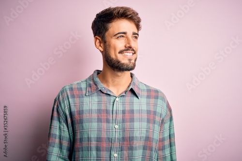 Young handsome man with beard wearing casual shirt standing over pink background looking away to side with smile on face, natural expression. Laughing confident.