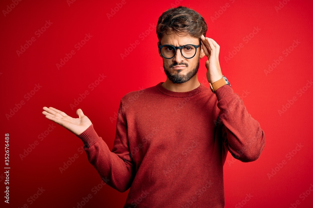 Young handsome man with beard wearing glasses and sweater standing over red background confused and annoyed with open palm showing copy space and pointing finger to forehead. Think about it. Young handsome man with beard wearing glasses and sweater standing over red background confused and annoyed with open palm showing copy space and pointing finger to forehead. Think about it.