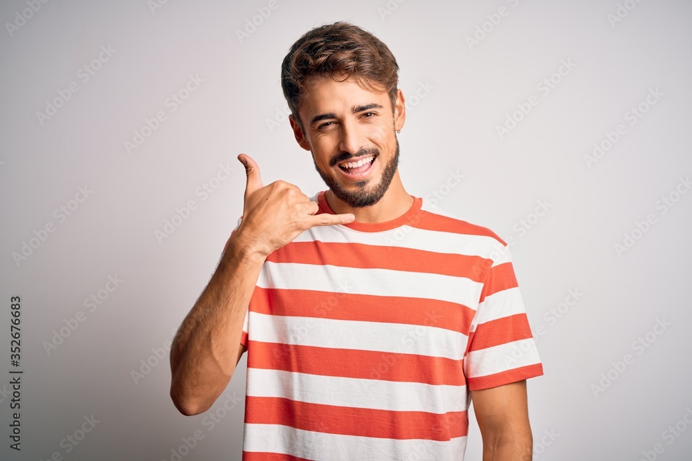 Young handsome man with beard wearing striped t-shirt standing over white background smiling doing phone gesture with hand and fingers like talking on the telephone. Communicating concepts. Young handsome man with beard wearing striped t-shirt standing over white background smiling doing phone gesture with hand and fingers like talking on the telephone. Communicating concepts.