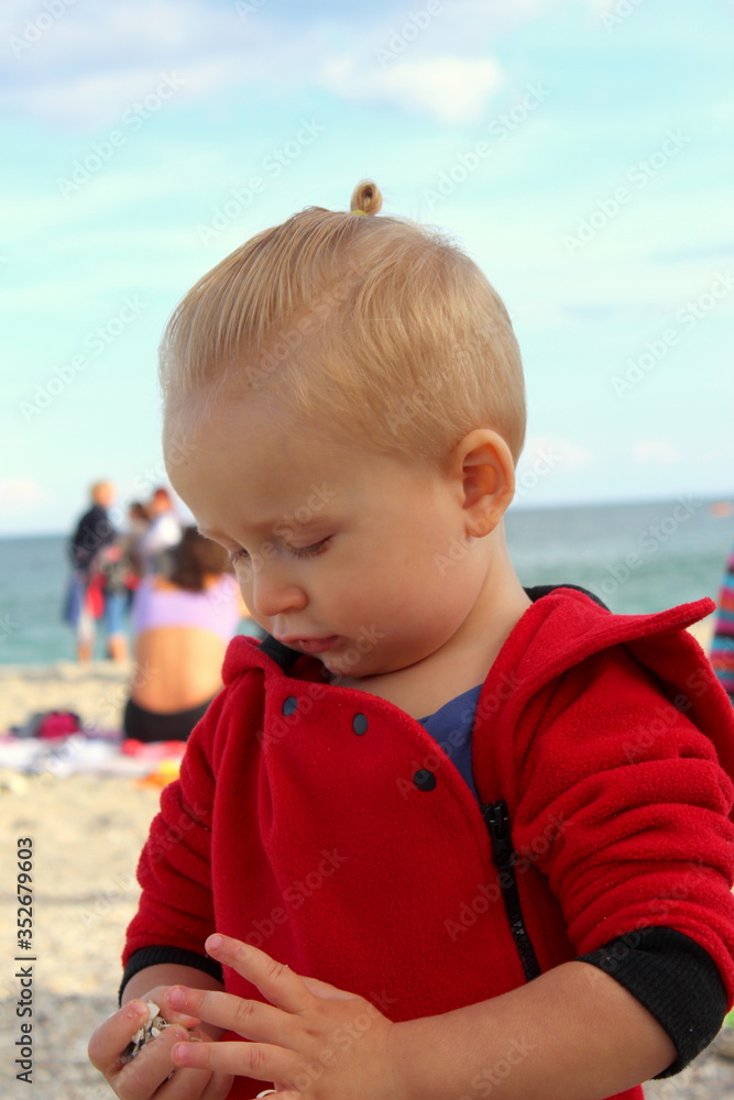 Little blond boy in red clothes stands on the sand on the shore by the sea in clear weather, selective focus. Little blond boy in red clothes stands on the sand on the shore by the sea in clear weather, selective focus.