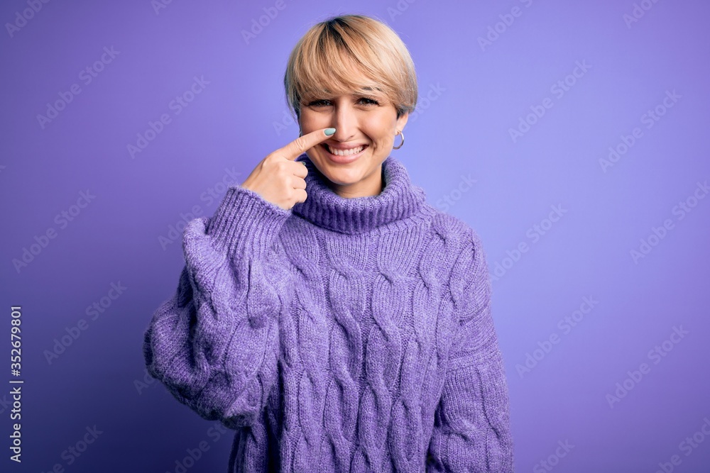 Young blonde woman with short hair wearing winter turtleneck sweater over purple background Pointing with hand finger to face and nose, smiling cheerful. Beauty concept Young blonde woman with short hair wearing winter turtleneck sweater over purple background Pointing with hand finger to face and nose, smiling cheerful. Beauty concept