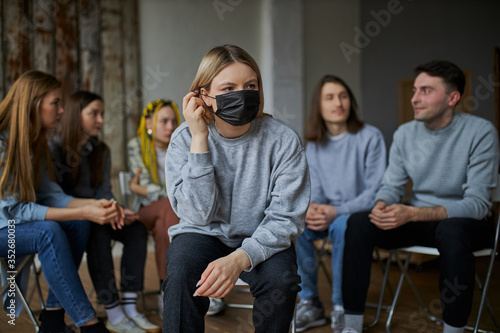 young caucasian female in mask suffering from coronavirus isolated in room with healthy people, sit keeping distance from group of people in the background