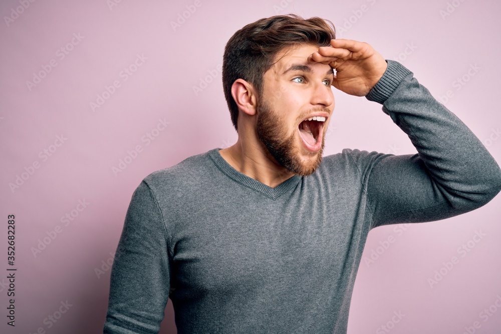 Young handsome blond man with beard and blue eyes wearing casual sweater very happy and smiling looking far away with hand over head. Searching concept. Young handsome blond man with beard and blue eyes wearing casual sweater very happy and smiling looking far away with hand over head. Searching concept.