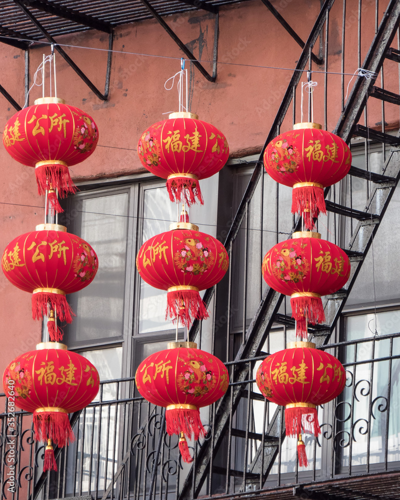 3 rows of Asian, bright, red paper lanterns hanging near a fire escape by apartments in a city.