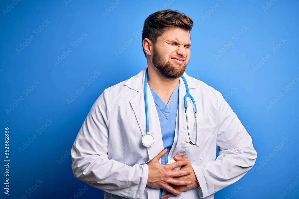 Young blond doctor man with beard and blue eyes wearing white coat and stethoscope with hand on stomach because nausea, painful disease feeling unwell. Ache concept. Young blond doctor man with beard and blue eyes wearing white coat and stethoscope with hand on stomach because nausea, painful disease feeling unwell. Ache concept.