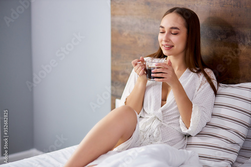 cute caucasian woman inhales the smell of coffee on bed, enjoy drinking coffee in the morning, wearing domestic bathrobe. relax, rest, weekends