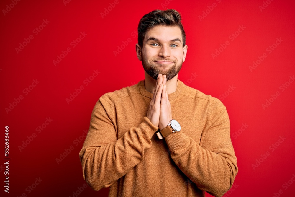 Young blond man with beard and blue eyes wearing casual shirt over red background praying with hands together asking for forgiveness smiling confident. Young blond man with beard and blue eyes wearing casual shirt over red background praying with hands together asking for forgiveness smiling confident.