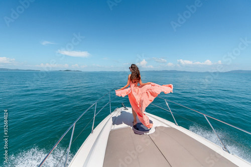 Outdoor shot of adorable young woman in a pink dress on edge of yacht, looking to beautiful nature landscape during trip. Happy woman enjoying summer travel. Back view