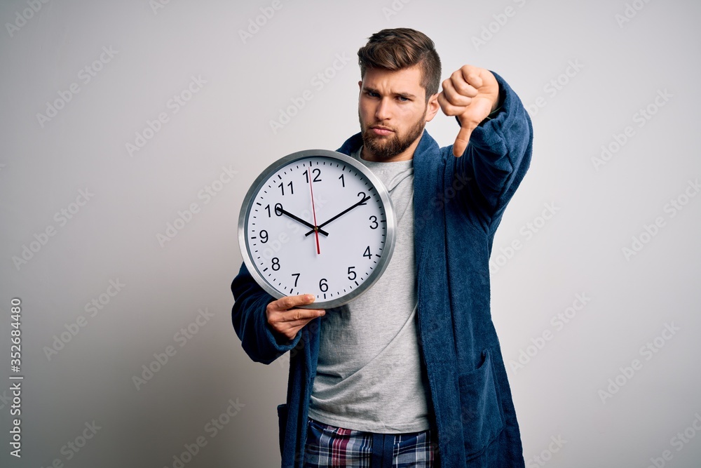 Young blond man with beard and blue eyes wearing pajama holding big clock with angry face, negative sign showing dislike with thumbs down, rejection concept Young blond man with beard and blue eyes wearing pajama holding big clock with angry face, negative sign showing dislike with thumbs down, rejection concept