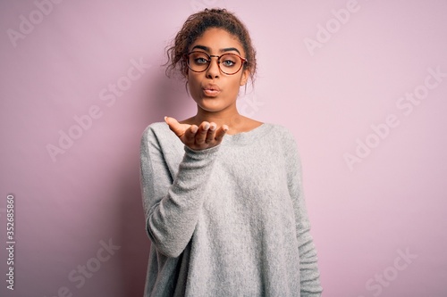 Young beautiful african american girl wearing sweater and glasses over pink background looking at the camera blowing a kiss with hand on air being lovely and sexy. Love expression.