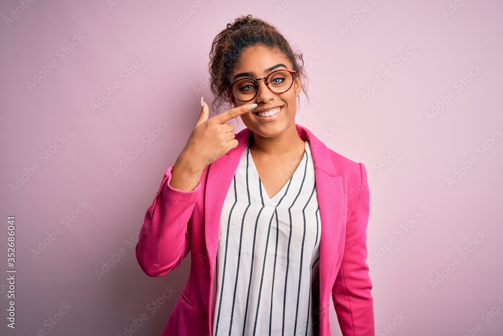 Beautiful african american businesswoman wearing jacket and glasses over pink background Pointing with hand finger to face and nose, smiling cheerful. Beauty concept