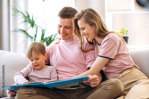 Family leisure at home. Young happy parents read a children's book with their adorable little son, sitting at home on the couch
