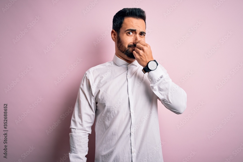 Young handsome man with beard wearing casual shirt standing over pink background smelling something stinky and disgusting, intolerable smell, holding breath with fingers on nose. Bad smell Young handsome man with beard wearing casual shirt standing over pink background smelling something stinky and disgusting, intolerable smell, holding breath with fingers on nose. Bad smell