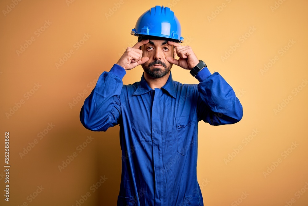 Mechanic man with beard wearing blue uniform and safety helmet over yellow background Trying to open eyes with fingers, sleepy and tired for morning fatigue
