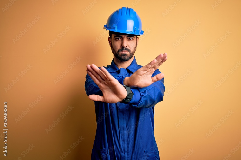 Mechanic man with beard wearing blue uniform and safety helmet over yellow background Rejection expression crossing arms doing negative sign, angry face