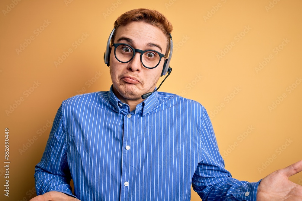 Fototapeta premium Young handsome redhead call center agent man wearing glasses working using headset clueless and confused expression with arms and hands raised. Doubt concept.
