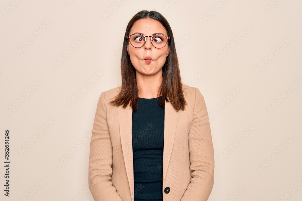 Young beautiful brunette businesswoman wearing jacket and glasses over white background making fish face with lips, crazy and comical gesture. Funny expression.