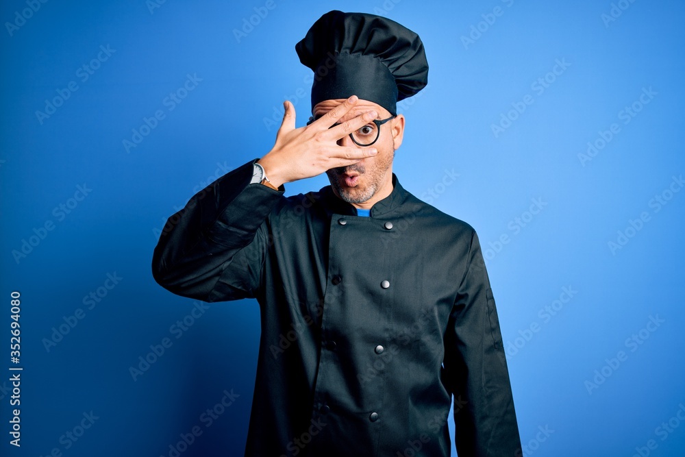 Young handsome chef man wearing cooker uniform and hat over isolated blue background peeking in shock covering face and eyes with hand, looking through fingers with embarrassed expression. Young handsome chef man wearing cooker uniform and hat over isolated blue background peeking in shock covering face and eyes with hand, looking through fingers with embarrassed expression.