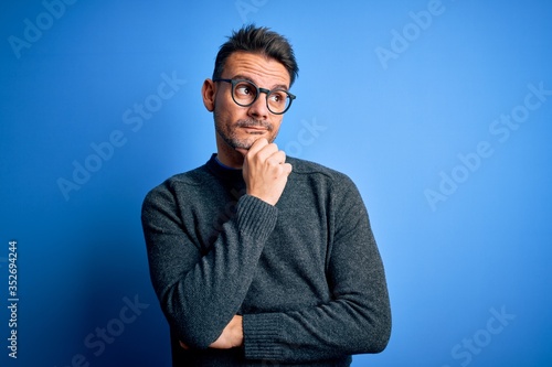 Young handsome man wearing casual sweater and glasses standing over blue background with hand on chin thinking about question, pensive expression. Smiling with thoughtful face. Doubt concept. © Krakenimages.com