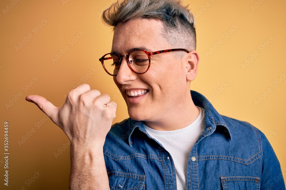 Close up of young handsome modern man wearing glasses and denim jacket over yellow background pointing and showing with thumb up to the side with happy face smiling Close up of young handsome modern man wearing glasses and denim jacket over yellow background pointing and showing with thumb up to the side with happy face smiling