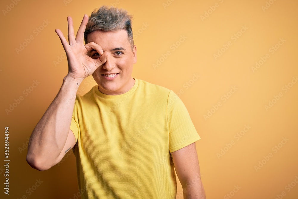 Young handsome modern man wearing yellow shirt over yellow isolated background doing ok gesture with hand smiling, eye looking through fingers with happy face. Young handsome modern man wearing yellow shirt over yellow isolated background doing ok gesture with hand smiling, eye looking through fingers with happy face.