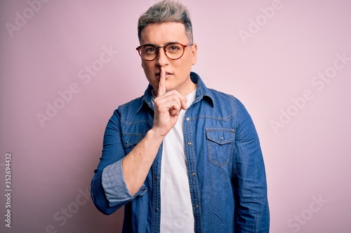 Young handsome modern man wearing glasses and denim jacket over pink isolated background asking to be quiet with finger on lips. Silence and secret concept.